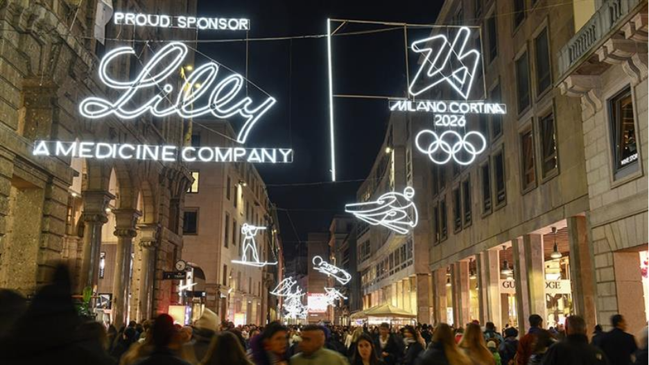 Nighttime view of a busy Milan shopping street illuminated with suspended light installations featuring winter sport figures, the Milano Cortina 2026 emblem and Olympic rings, and prominent &ldquo;Lilly &ndash; A Medicine Company&rdquo; sponsor signage glowing above the crowd.